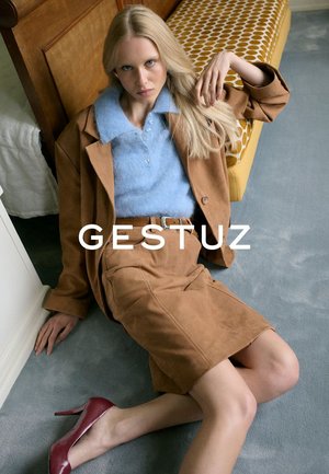 Bruine suède blazer en rok met een lichtblauwe sweater; bordeaux hoge hakken; binnenomgeving met gestoffeerde gele patronen.