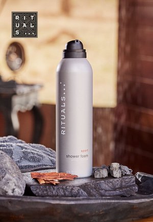 Gray metal canister of shower foam with black nozzle, labeled "RITUALS sport shower foam," placed on textured stones in a natural setting.