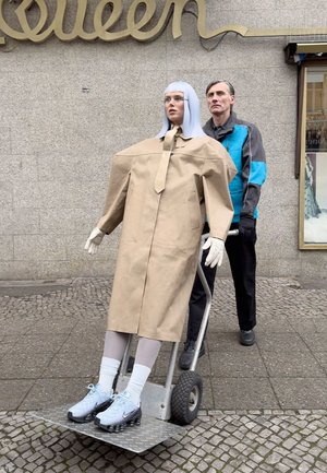 Schaufensterpuppe trägt einen beige übergroßen Mantel und eine Krawatte, weiße Leggings und hellblaue Schuhe und wird von einer Person in einer blauen Jacke auf einem Wagen geschoben.
