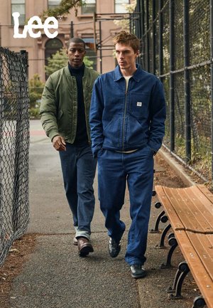Dark denim jacket with blue jeans on one person; green bomber jacket and navy sweater on the other. Outdoors, casual setting with a bench.