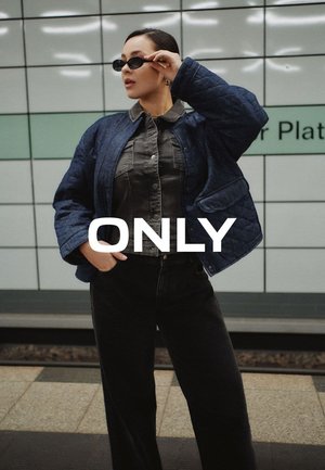 Donna con occhiali da sole, giacca trapuntata blu e pantaloni scuri in piedi in una stazione della metropolitana con la parola "ONLY" sovrapposta.