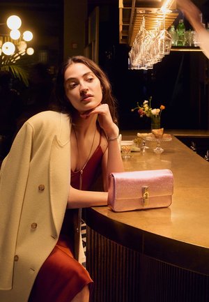 Metaliczna różowa kopertówka z zapięciem, leżąca na blacie z złotego pręta. Miękki żółty płaszcz i głęboka czerwona sukienka noszone przez siedzącą postać.