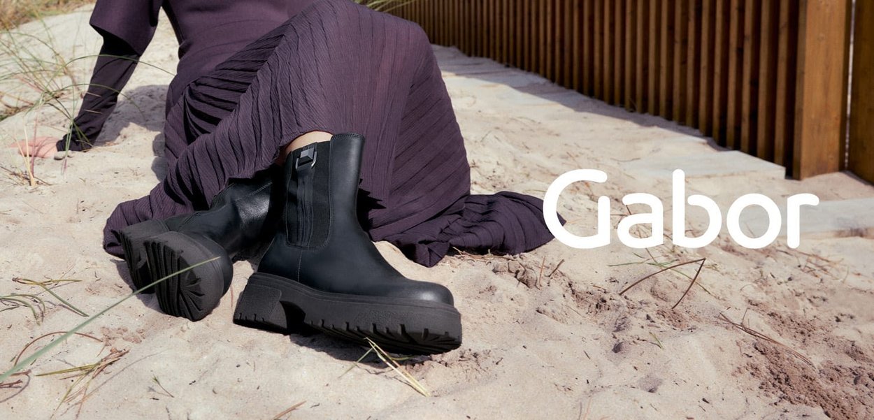Person, der auf dem sandigen Boden sitzt und schwarze klobige Stiefel sowie einen plissierten dunkelvioletten Rock trägt, in der Nähe eines Holzzauns.
