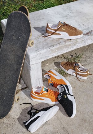 Patineta apoyada en un banco de piedra con zapatillas Puma marrones, naranjas y negras esparcidas sobre y cerca del banco al aire libre sobre hormigón y césped.