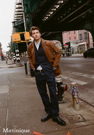 Brauner Mantel über einer marineblauen Weste, weißem Hemd und dunklen Jeans. Schwarze Stiefel. Urbane Umgebung mit Graffiti und einer Hochbahn im Hintergrund.