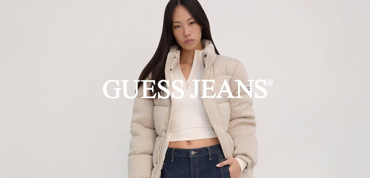 Beige pufferjack met een hoge kraag, ritssluiting aan de voorkant en grote gequiltte secties, gecombineerd met een witte aangesloten top en donkere jeans.