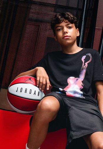 Schwarzes T-Shirt mit einem bunten Grafikeindruck eines Basketballspielers, kombiniert mit schwarzen Shorts und einem rot-weißen Basketball, der mit "JORDAN" beschriftet ist.