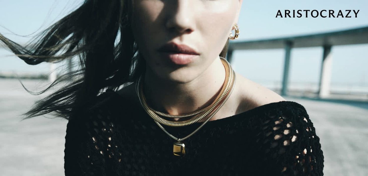 Collares en tono dorado con capas y un colgante cuadrado en una modelo que lleva una blusa de punto negra, con cabello suelto y un fondo minimalista.