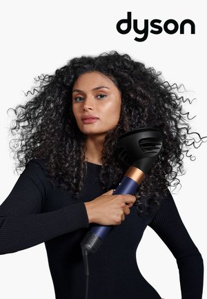 Donna con capelli ricci che utilizza un diffusore per asciugacapelli Dyson, indossando una maglietta nera a maniche lunghe, su uno sfondo bianco con il logo Dyson.