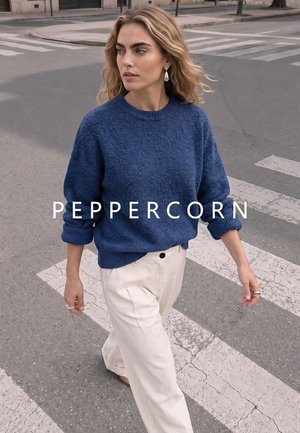 Jonge vrouw in blauwe trui en witte broek steekt een stadsvoetgangersoversteekplaats over met tekst "PEPPERCORN" eroverheen geplaatst.