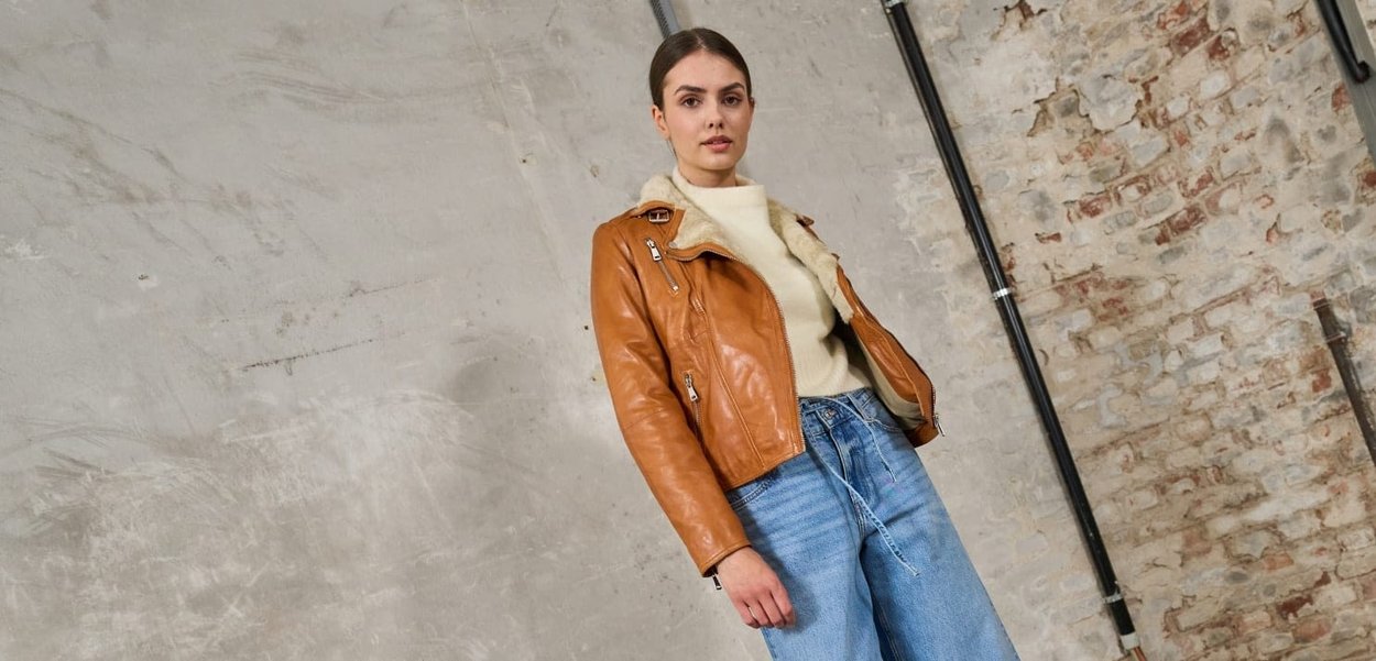Tan leren bikerjack met schapenvacht kraag, ritszakken, gecombineerd met lichtblauwe, ruime jeans. Neutrale trui eronder.