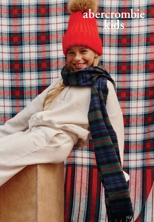 Czerwona dzianinowa czapka z pomponem, kremowa bluza i spodnie, szal w niebiesko-zieloną kratę. Tło zdobi wzór w czerwoną i niebieską kratę.