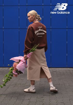 New Balance-sweatshirt in donkerbruin, beige rok met zijsplit, witte schoenen, houdt roze bloemen vast die in papier zijn gewikkeld. Blauwe achtergrond.