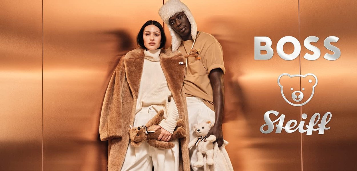 Zwei Models stehen vor einem kupferfarbenen Hintergrund. Die Frau trägt einen langen, braunen Kunstpelzmantel und einen weißen Pullover und hält einen Teddybär. Der Mann trägt ein beiges Hemd mit einem Teddybär-Print und hält einen weißen Teddybären.