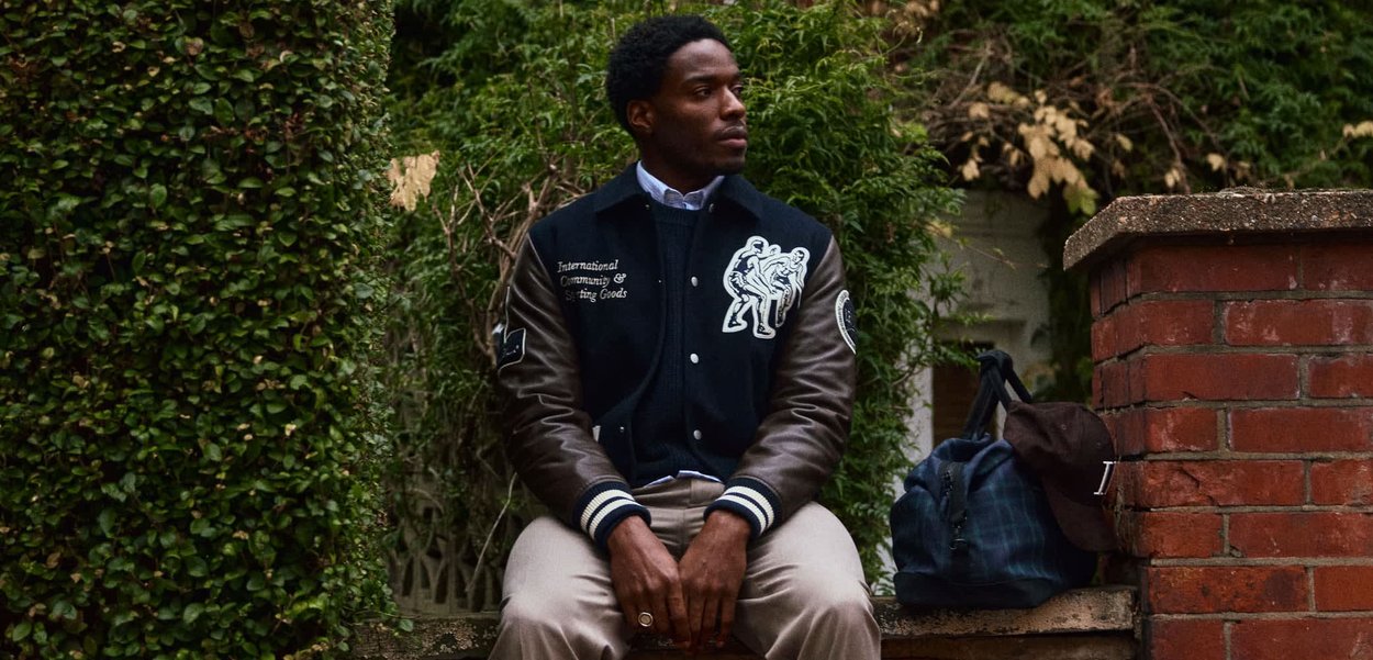 Mand iført en sort varsityjakke og beige bukser, siddende på en stenskrænt ved siden af en blå taske og en brun kasket, omgivet af grønt område.