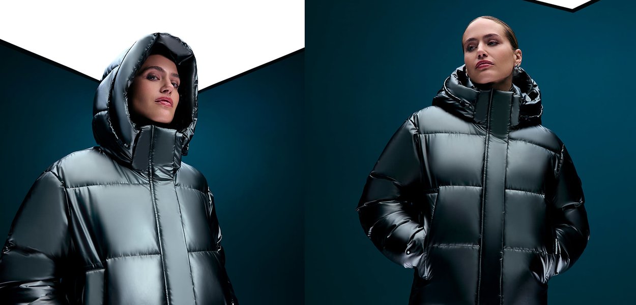 Nainen, jolla on päällä kiiltävä musta toppatakki, huppu päässä, kädet taskuissa, katsoo hieman sivulle tumman tealinsinistä taustaa vasten.