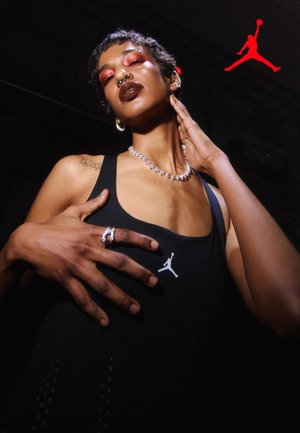Person mit kurzen Haaren und auffälligem Make-up, die ein schwarzes ärmelloses Oberteil mit weißem Jordan-Logo trägt, berührt Hals und Brust, hervorgehoben durch einen dunklen Hintergrund.