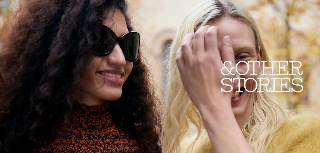 To kvinder, der smiler udendørs; den ene med krøllet mørkt hår og solbriller, den anden med glat blondt hår, der justerer sit hår.