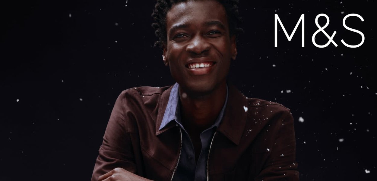 Bruine jas met een textuurafwerking, over een blauw hemd gedragen. De achtergrond is donker met witte stippen die op sneeuwvlokken lijken. M&S-logo zichtbaar.