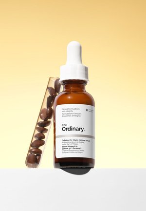 Botella de cristal marrón con gotero etiquetada como The Ordinary Caffeine 3% + Escin 1% suero facial junto a un tubo de ensayo con granos de café marrones.
