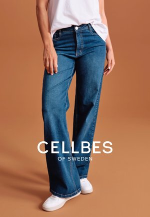Blue denim wide-leg pants with a slight fade, five-pocket design, and a classic fit. Styled with white sneakers on a beige background.