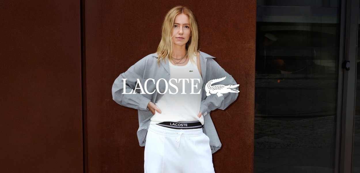 Giovane donna con capelli biondi che indossa abbigliamento sportivo bianco Lacoste e giacca leggera, in piedi con le mani sui fianchi contro un muro marrone.
