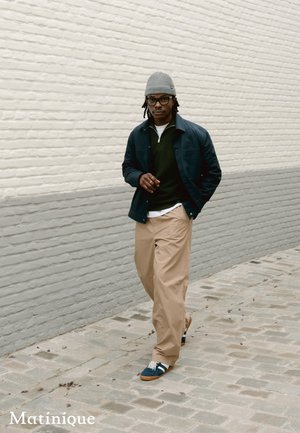 Mann, der auf einer Kopfsteinpflasterstraße geht und eine graue Mütze, eine Brille, eine marineblaue Jacke, einen dunkelgrünen Pullover, beige Hose und blaue Turnschuhe trägt.