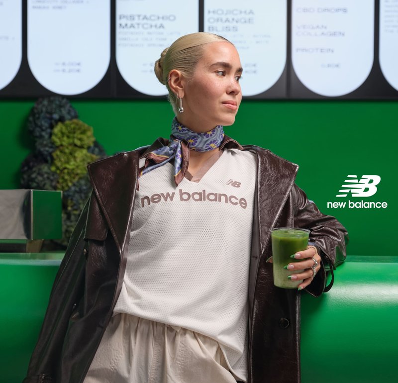 Giovane donna con abbigliamento sportivo New Balance e giacca di pelle tiene un frullato verde seduta di fronte a un muro verde con insegne del menù.