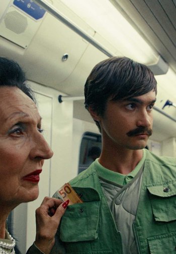 Due persone in una metropolitana: una donna più anziana con i capelli scuri e rossetto rosso, e un giovane in una giacca verde chiaro, che tiene in mano una banconota da 50 euro.