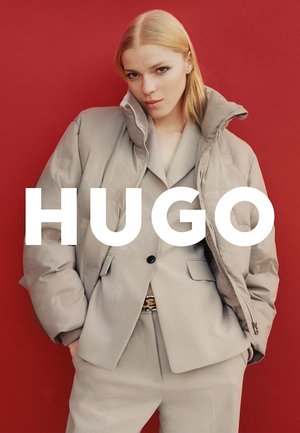 Giacca imbottita color beige chiaro con collo alto sopra un completo coordinato. Texture liscia, chiusura con cerniera e motivo logo sottile. Sfondo rosso intenso.