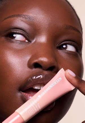 Lip treatment tube with pink packaging, matte finish, and white text held by a model with smooth, glistening skin and an open mouth.