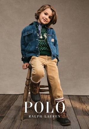 Veste en denim, pull à motifs verts, pantalons en velours côtelé beige, bottes marron. Enfant assis sur un tabouret en bois avec un fond neutre.