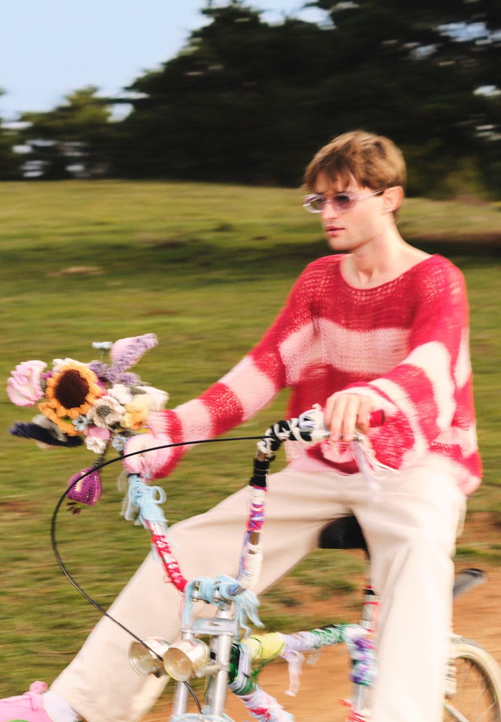 Maglione a righe rosse e bianche, pantaloni beige; bicicletta decorata con filato colorato e un mazzo di fiori sul manubrio.