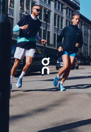 Deux coureurs à l'extérieur. L'homme porte un haut bleu marine à manches courtes avec un design dégradé, un short blanc et des chaussures bleues. La femme porte une veste noire, un short bleu et des chaussures bleues.
