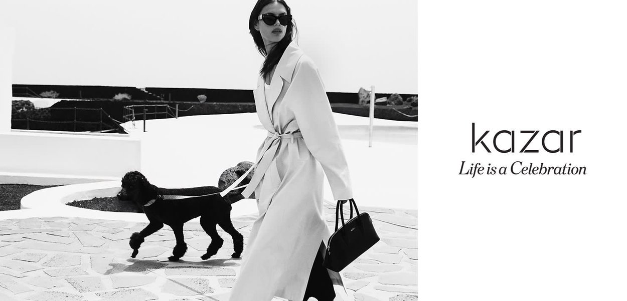 Black and white image of a woman in a long, light-colored coat walking a black dog. She holds a black handbag and wears sunglasses.