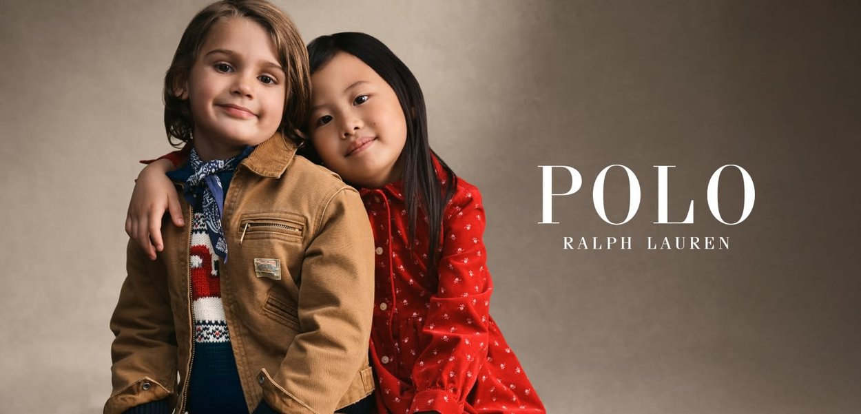 To børn står tæt sammen. Drengen har en beige jakke på over en mønstret sweater, og pigen har en rød kjole med prikker på.