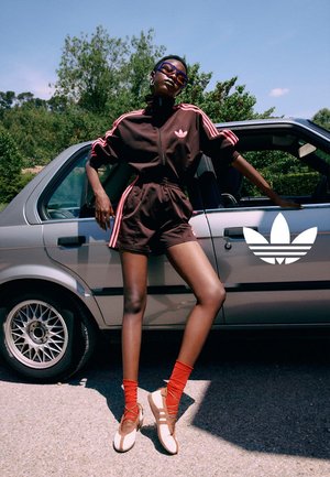 Persona con tuta Adidas scura e calzini rossi si appoggia a un'auto argento con logo Adidas sulla porta, all'aperto con alberi sullo sfondo.