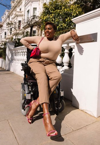 En kvinna i rullstol bär en beige ribbad topp, beige byxor och rosa sandaler med remmar, och har en liten rosa handväska hängande på armen.
