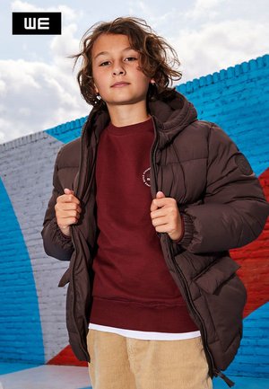 Brun oppustelig jakke med hætte, med lynlås og ribbede manchetter, lagd over en burgunder sweatshirt. Modellen har beige fløjl bukser på.