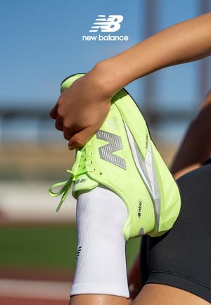 Neon yellow athletic shoe with silver accents and textured design, being held by a hand. White sock visible, background out of focus.