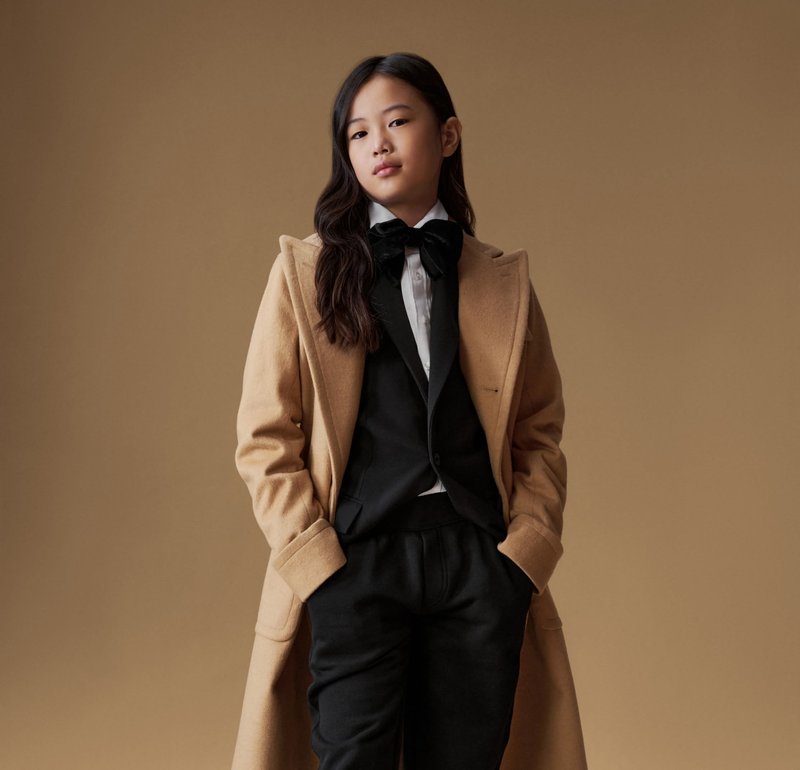 Manteau en laine tan sur un costume noir avec une chemise blanche et un nœud papillon. Présente un design épuré, une texture lisse et une coupe ajustée.
