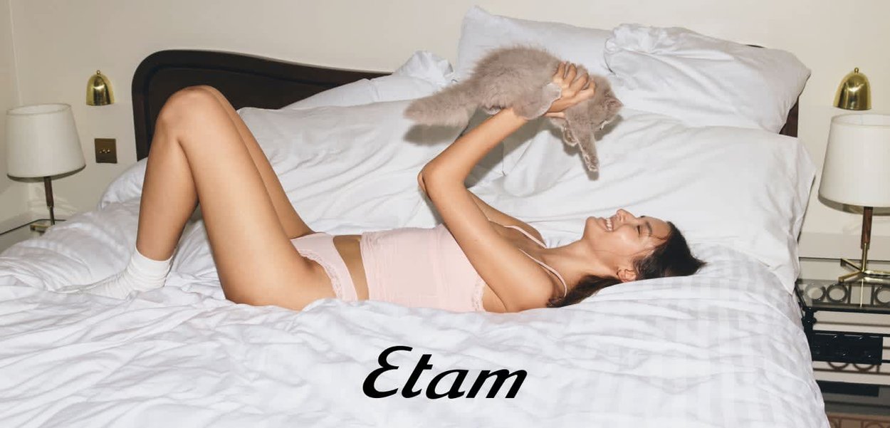 Frau in hellrosa Unterwäsche und weißen Socken liegt auf dem Bett, hält eine flauschige graue Katze hoch und lächelt, mit weißen Kissen und Lampen im Hintergrund.