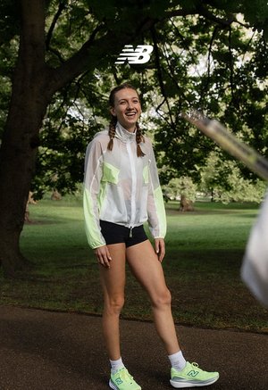 Kobieta uśmiechająca się na zewnątrz na ścieżce w parku z drzewami w tle, ubrana w neonowo-zielone buty do biegania New Balance i odzież sportową.