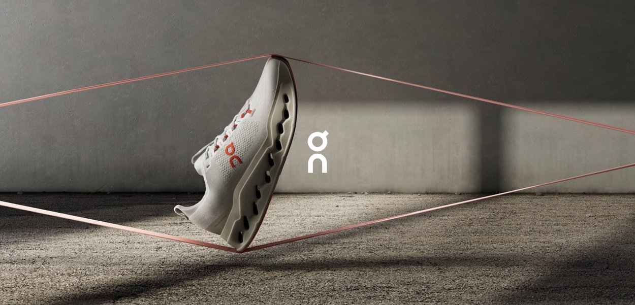 White athletic shoe with textured knit upper, orange accents, and a unique wavy sole design. Background features concrete and pink lines.