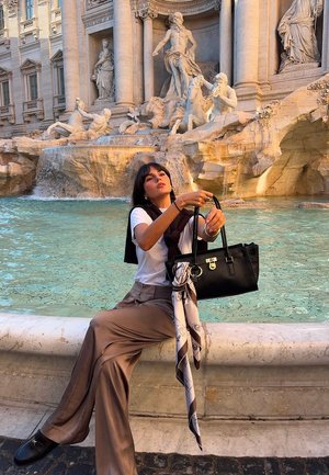 Donna con camicia bianca e pantaloni marroni seduta presso la Fontana di Trevi, mentre tiene una borsa nera con una sciarpa decorata annodata.