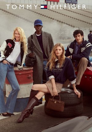 Group of four people in casual clothing, featuring a black varsity jacket, a grey coat, a navy sweatshirt, and a plaid skirt. Background includes tires and a racetrack.