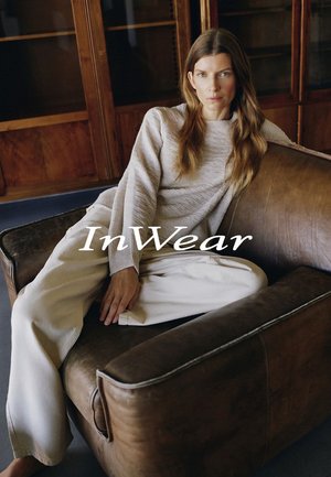 Frau mit langen Haaren in beigefarbenem Pullover und weißer Hose lehnt auf einem braunen Ledersessel in einem holzvertäfelten Raum, der Schriftzug "InWear" ist sichtbar.