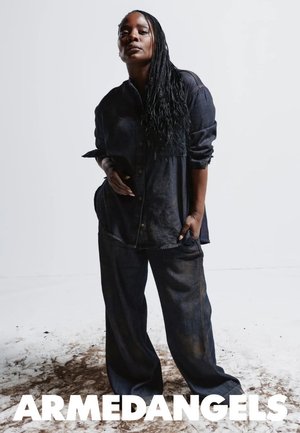 Zelfverzekerd persoon met lang gevlochten haar draagt loszittend donker denim overhemd en broek, staand op een met vuil besmeurde vloer tegen een effen witte achtergrond.