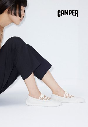 Kobieta z krótkimi czarnymi włosami siedzi bokiem, ubrana w czarne spodnie i białe wsuwane buty na sznurowadła na jednolitym białym tle.