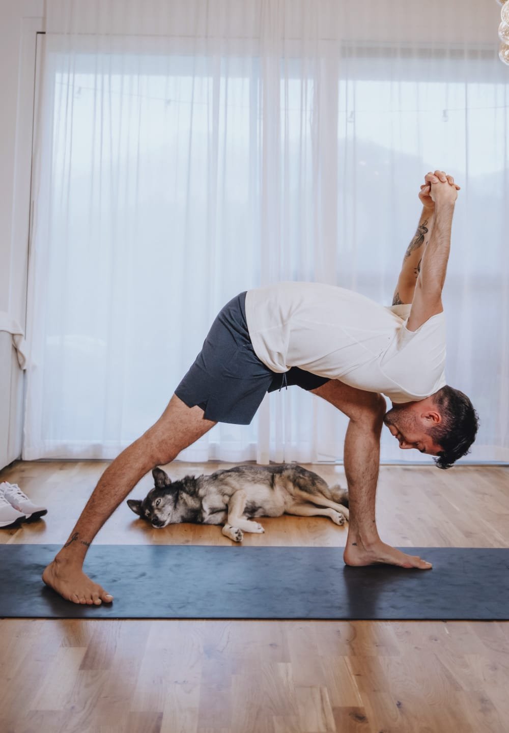 Person practicing yoga on a black mat, wearing a white shirt and gray shorts. A dog rests on a wooden floor nearby, with sheer curtains in the background.